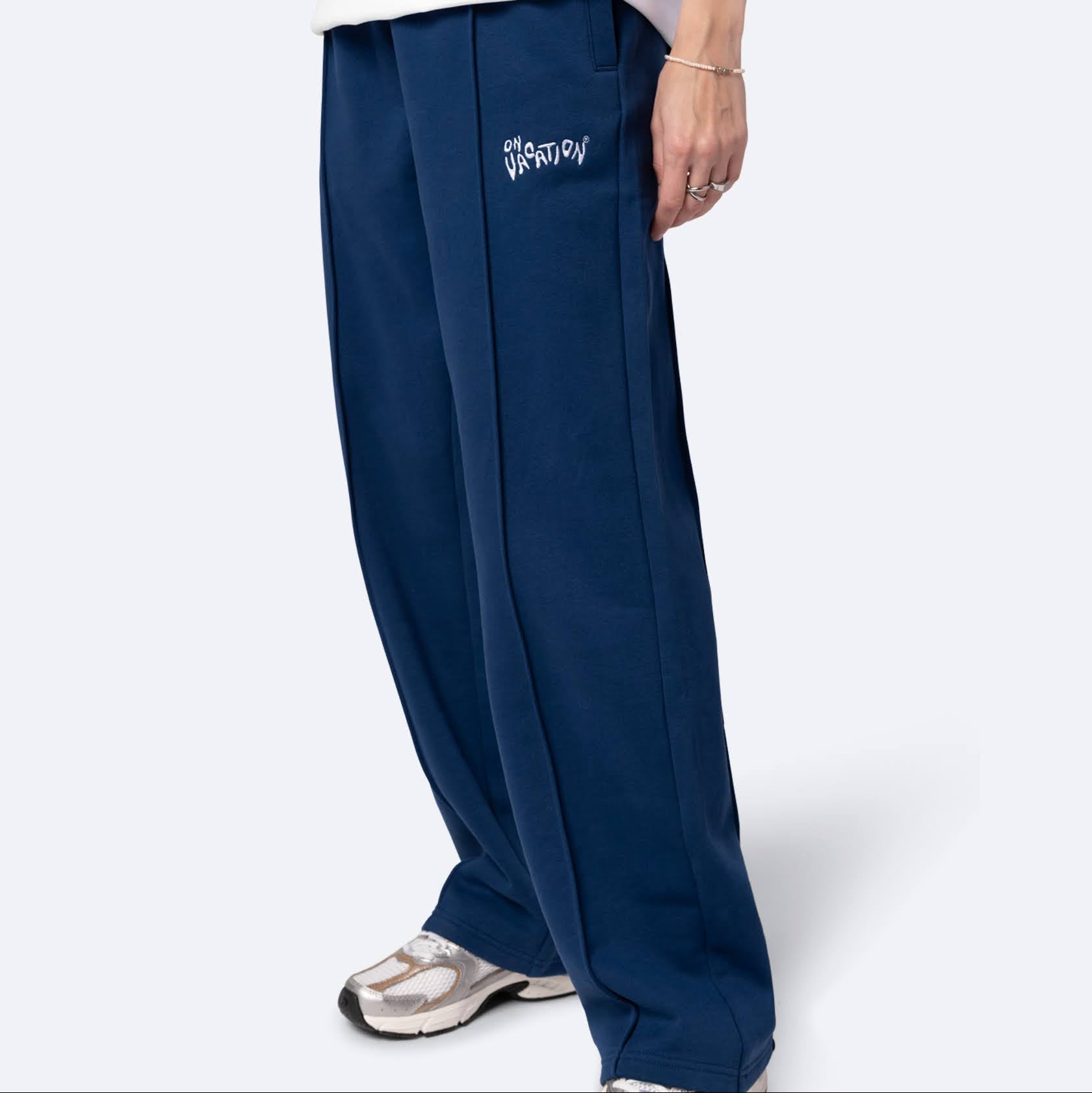 Eine Person, die eine marineblaue Hose mit weitem Bein und weißem Schriftzug, ein weißes T-Shirt, weiße Turnschuhe und ein silbernes Armband trägt, steht vor einem schlichten hellen Hintergrund.