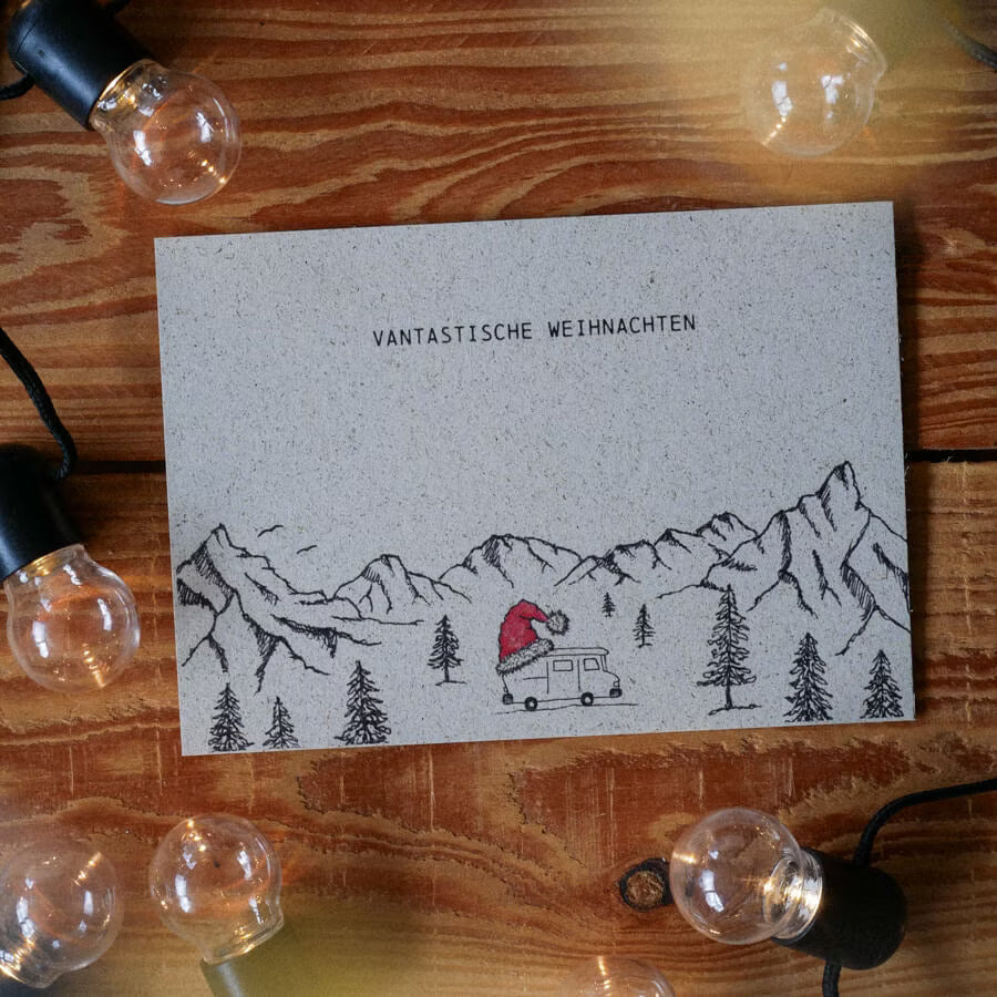 Graue Postkarte mit gezeichneten Bergen, Bäumen und einem Wohnwagen. Text: "Vantastische Weihnachten". Lichterkette daneben.