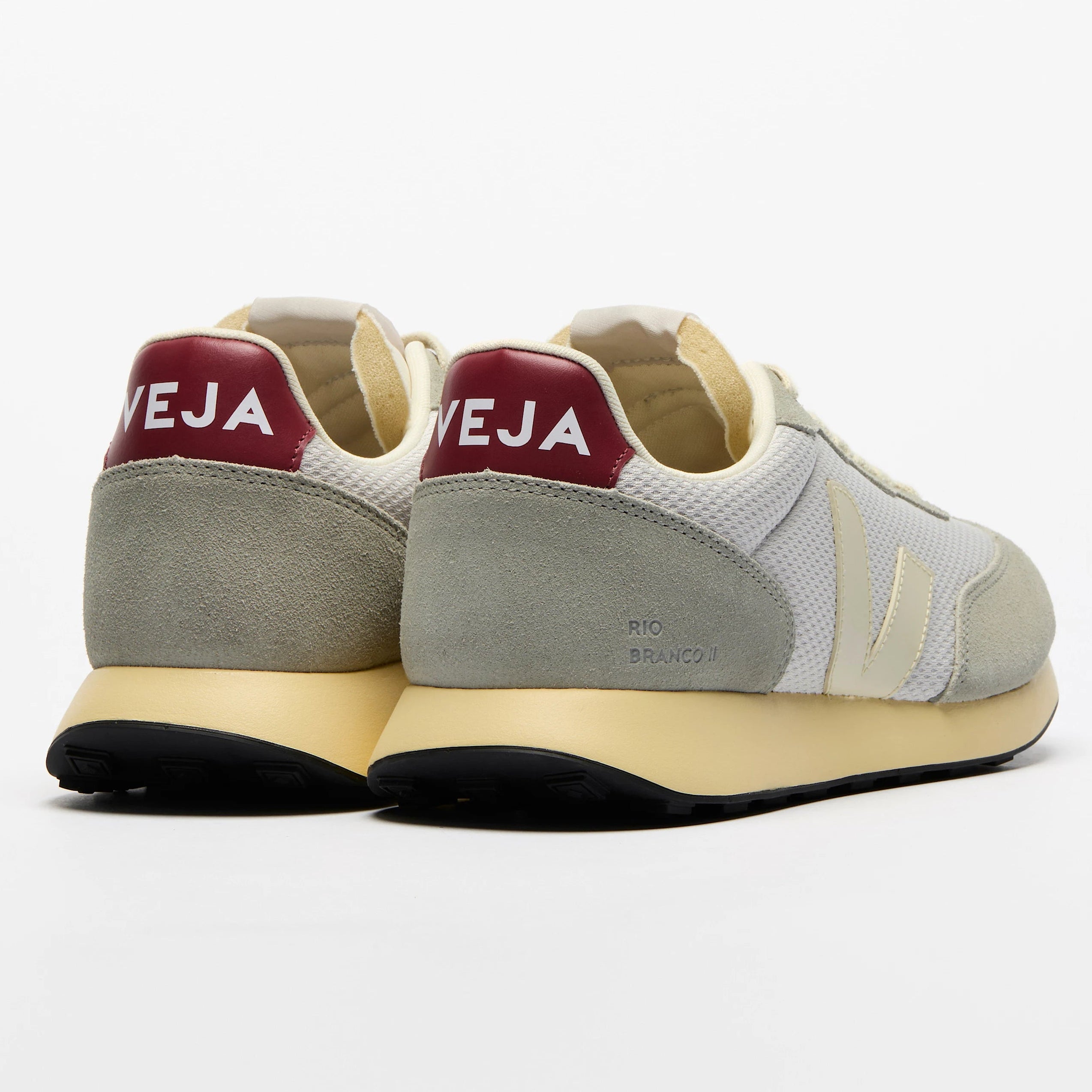 Ein Paar Veja Sneakers in Hellgrau und Weiß mit beiger Sohle, von hinten gesehen. An der Ferse befindet sich ein weinrotes Feld mit weißer Schrift VEJA. Die Schuhe sind vor einem schlichten weißen Hintergrund platziert.