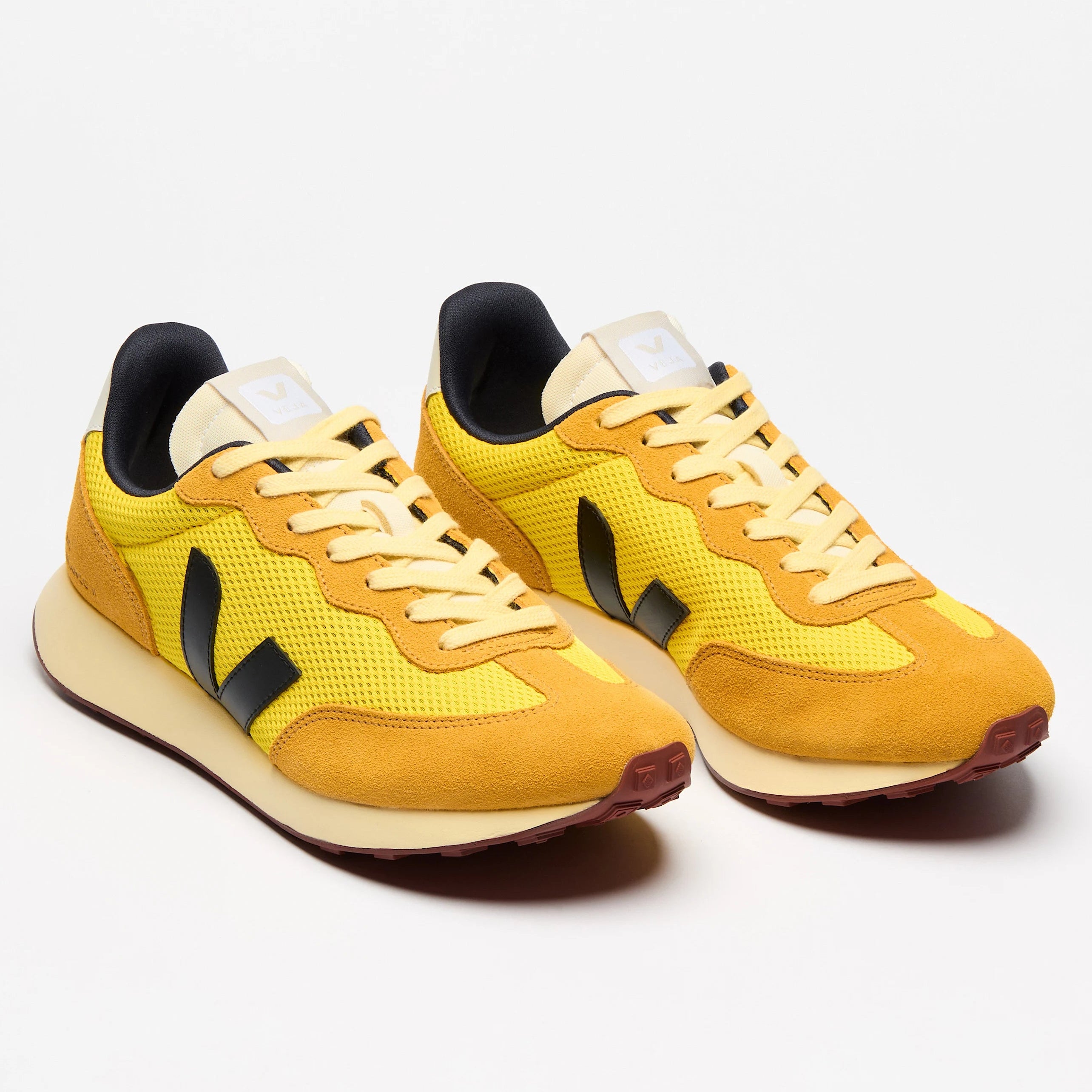 Ein Paar gelbe Turnschuhe mit schwarzen Akzenten und weißen Sohlen, aus einem leichten Winkel vor einem schlichten weißen Hintergrund betrachtet. Die Schuhe haben ein sportliches, retro-inspiriertes Design mit Schnürsenkeln und einem Logo an der Seite.