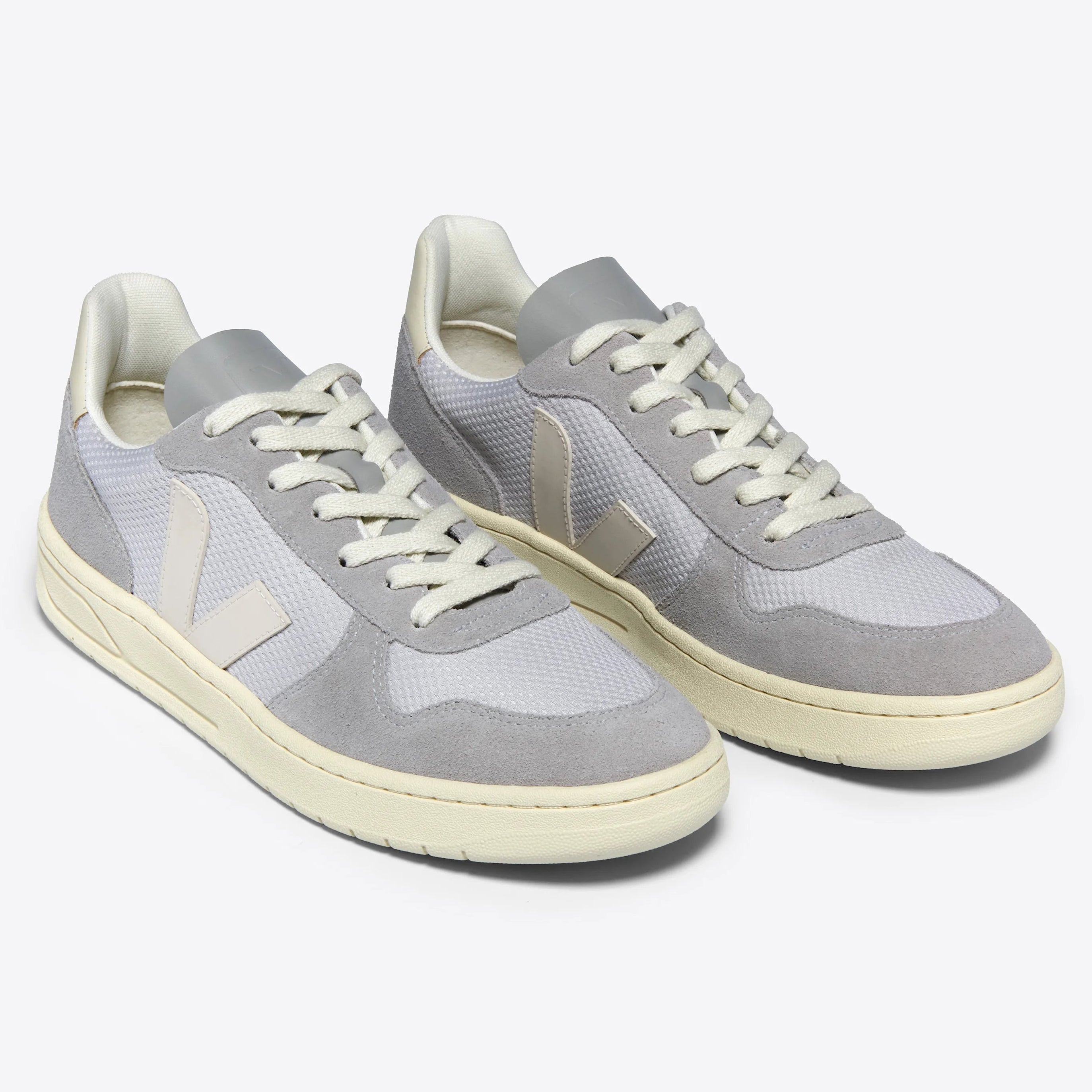 Ein Paar hellgraue und weiße Sneaker mit beiger Sohle, weißen Schnürsenkeln und einem Wildleder-Finish an der Spitze und den Seiten, mit einem stilisierten V-Logo an der Seite.