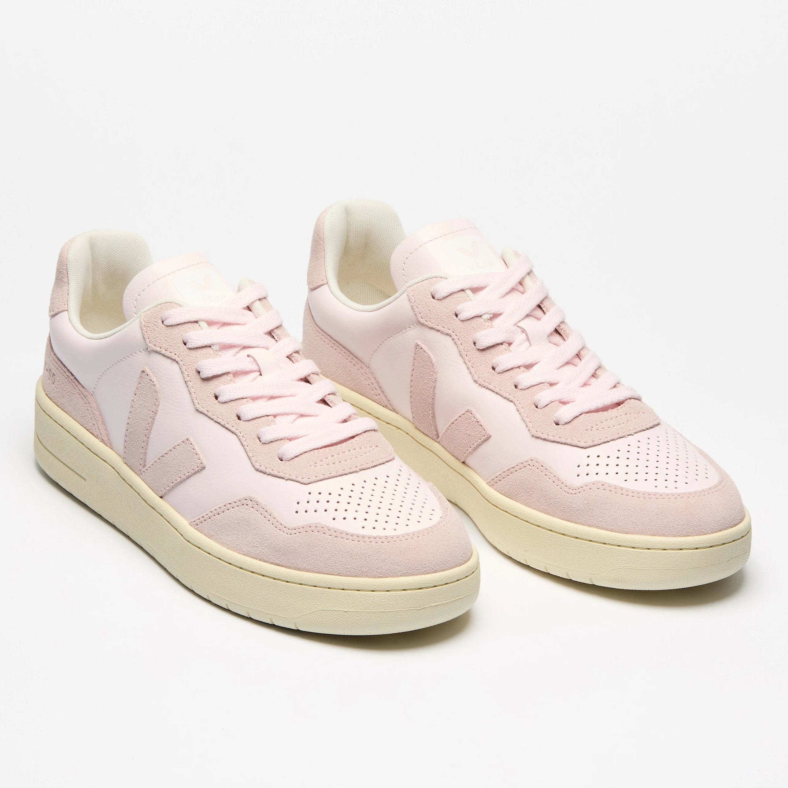 Ein Paar hellrosa und cremefarbene Turnschuhe mit weißer Sohle, mit perforierten Details an der Spitze und an den Seiten, auf einem einfarbig weißen Hintergrund.