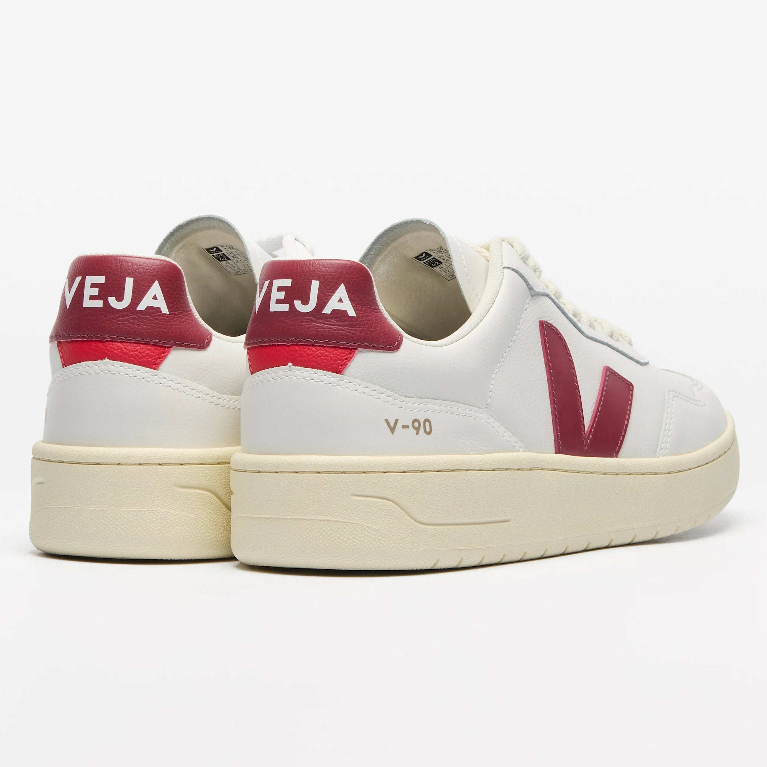 Ein Paar weiße Turnschuhe mit roten Fersenakzenten, rotem VEJA-Branding auf der Rückseite, roten V-Logos an den Seiten und cremefarbenen Sohlen. Die Schuhe sind so positioniert, dass die Fersen nach vorne zeigen.