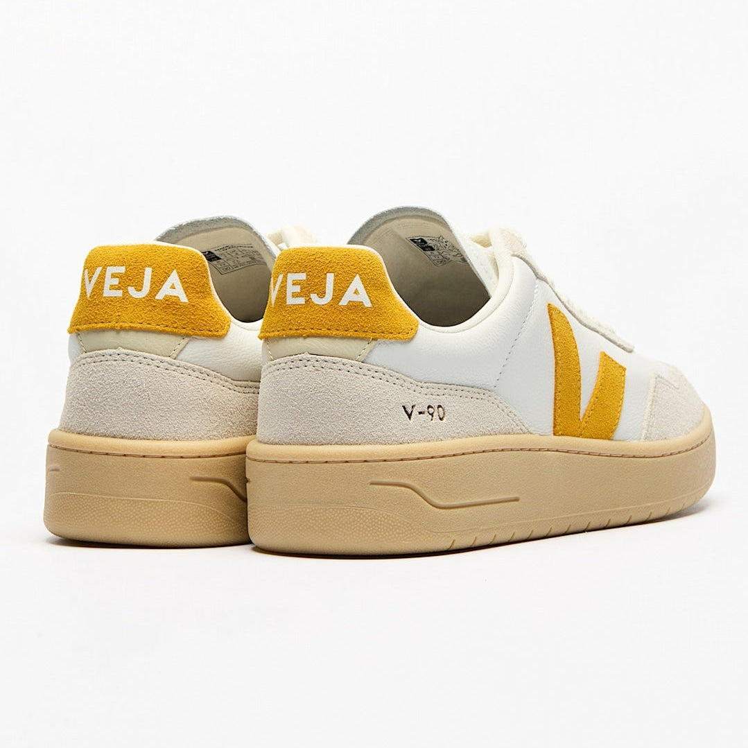Ein Paar weiße Veja V-90 Sneakers mit gelben Akzenten, einschließlich der Fersenlasche mit VEJA-Text und seitlichem Logo, und beigen Sohlen, von hinten und von der Seite auf weißem Hintergrund dargestellt.