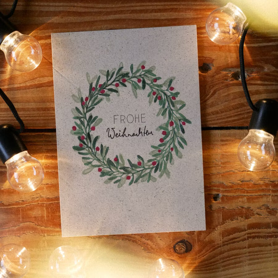Weihnachtskarte mit grünem Kranz und "Frohe Weihnachten", umgeben von Lichterkette auf einem Holztisch.