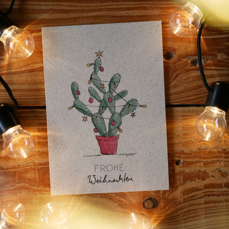Karte mit einem gezeichneten Kaktus als Weihnachtsbaum, umrahmt von Lichterketten, auf einem Holztisch.