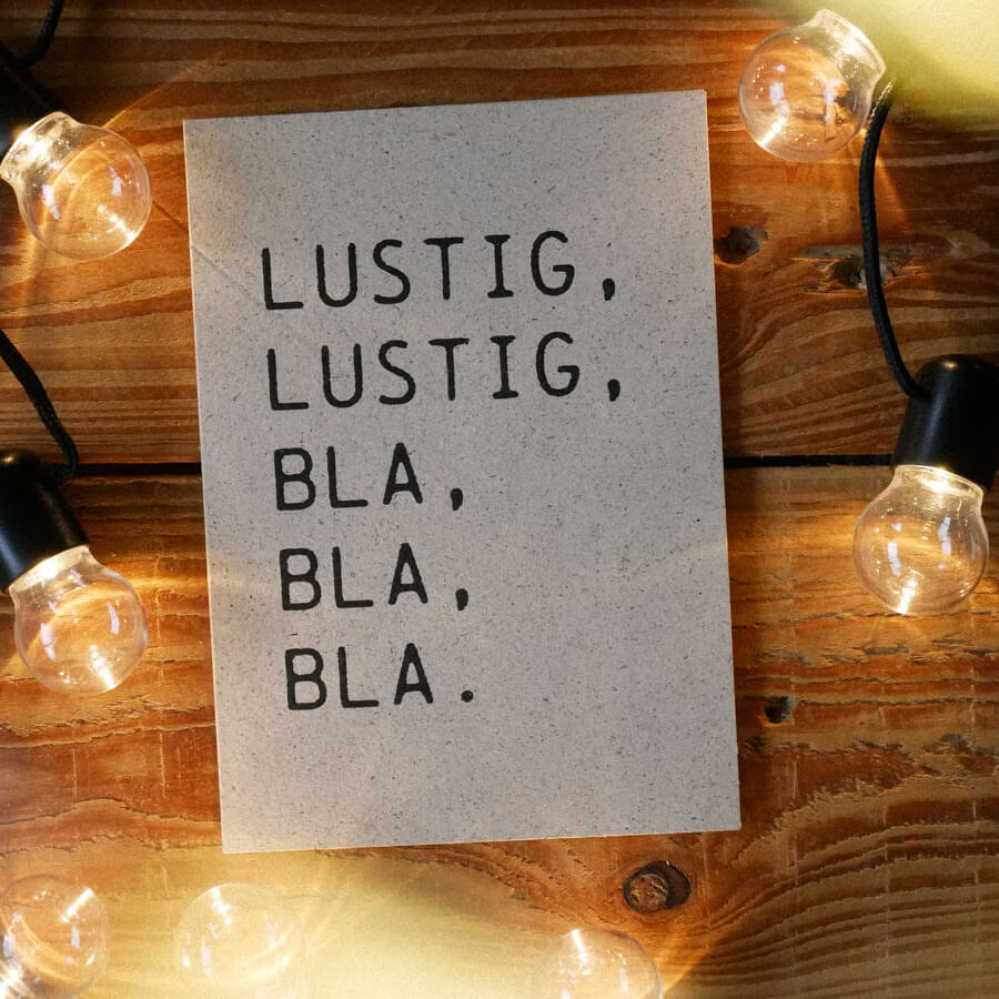 Eine Karte mit der Aufschrift "LUSTIG, LUSTIG, BLA, BLA, BLA" liegt auf einem Holztisch, umgeben von Glühbirnen.