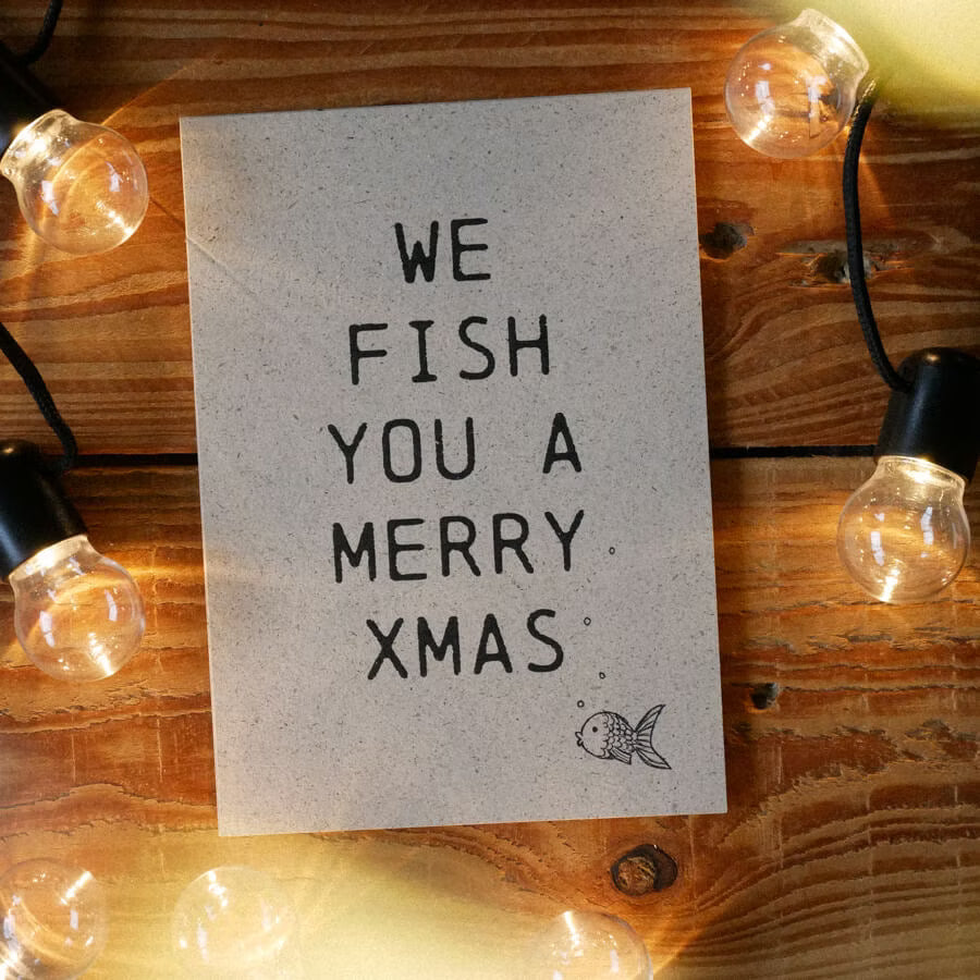 Karte mit dem Text "WE FISH YOU A MERRY XMAS" und einem kleinen gezeichneten Fisch, umgeben von Lichterketten.