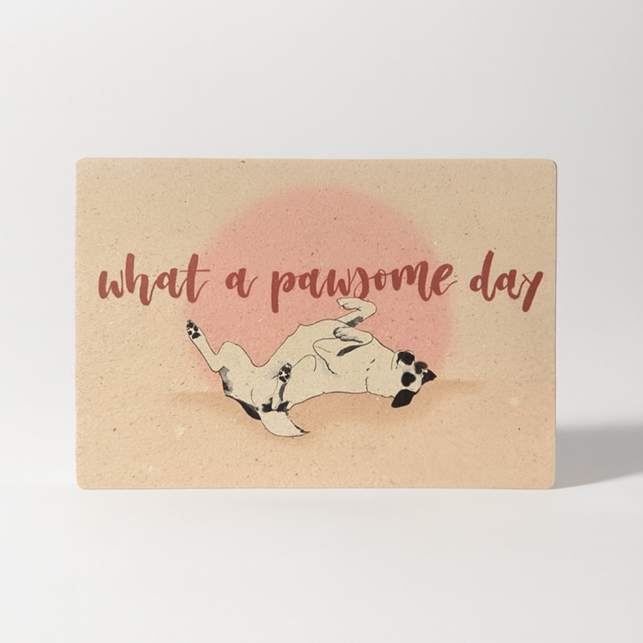 Eine Karte mit dem Text "What a pawsome day" über einer verspielten Illustration eines auf dem Rücken liegenden Hundes vor einer rosa Sonne auf beigem Hintergrund.