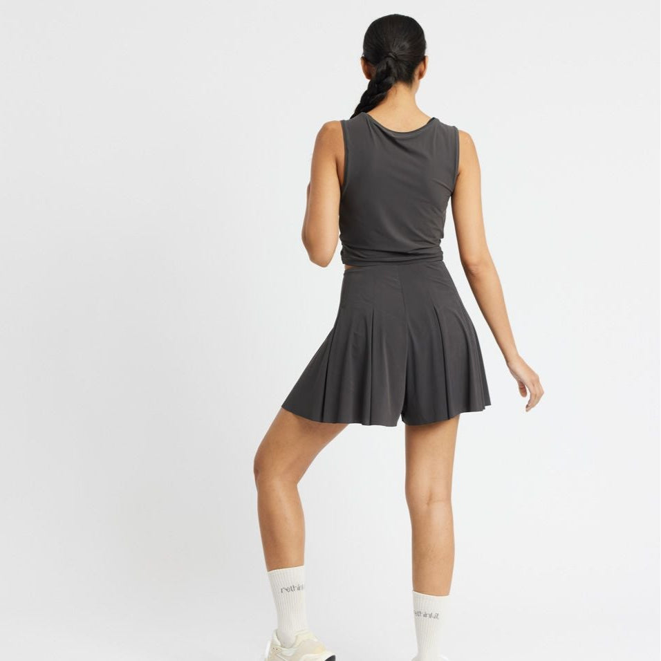 Person in a black sleeveless top and pleated skirt, standing with back turned, wearing socks and sneakers.