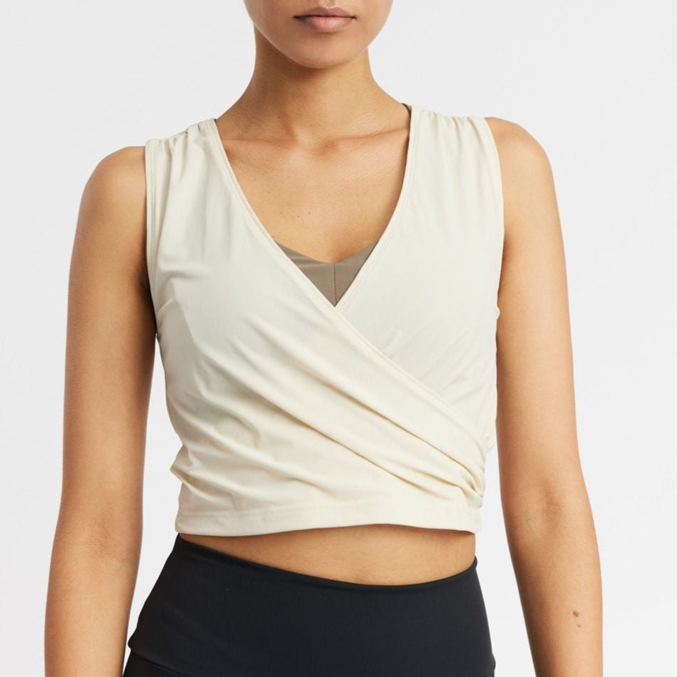 Person in a sleeveless, cream-colored wrap top and black pants, shown from the waist up against a white background.