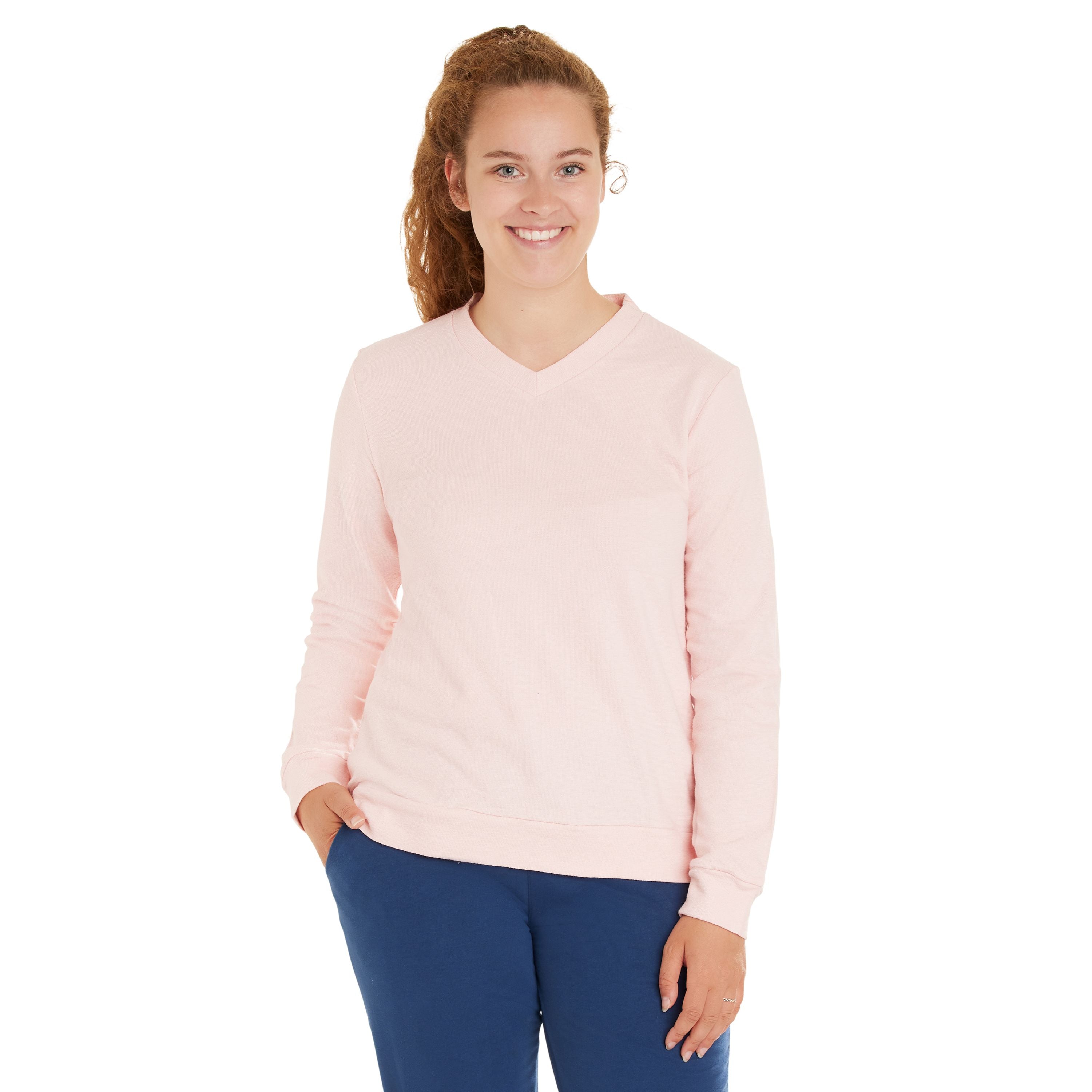 Eine Frau mit langen lockigen Haaren, die einen hellrosa Pullover mit V-Ausschnitt und eine blaue Hose trägt, steht lächelnd mit einer Hand in der Tasche vor einem weißen Hintergrund.