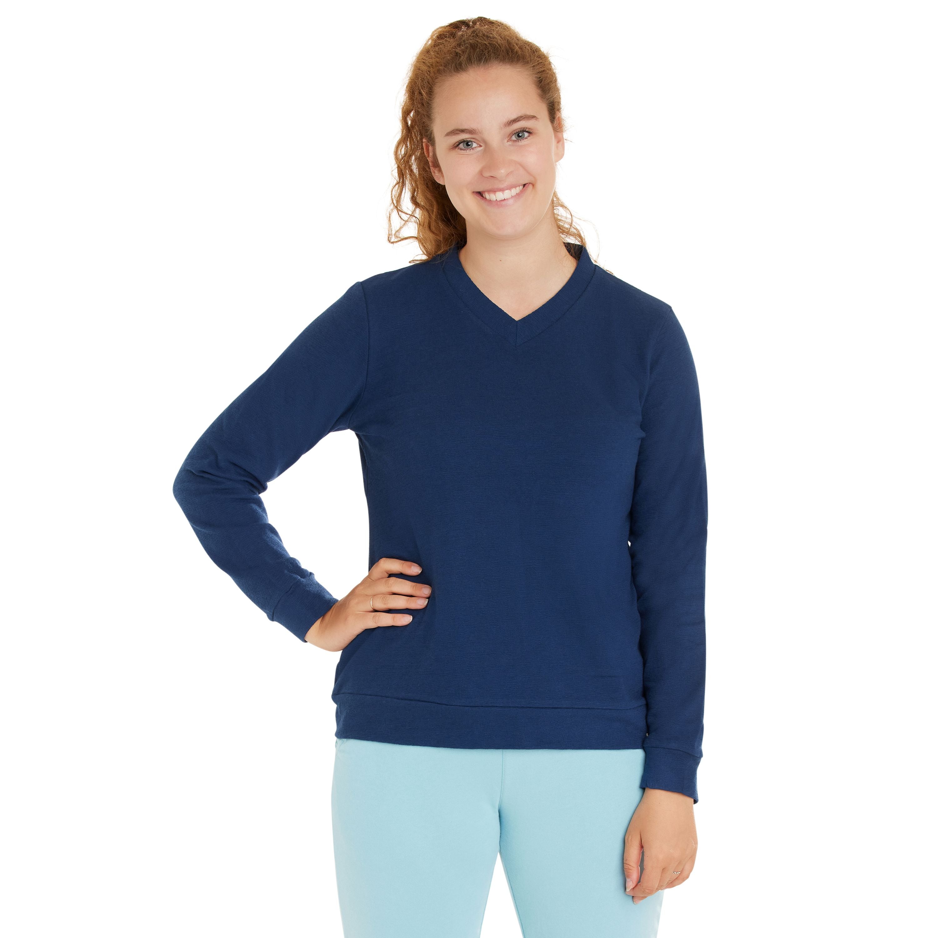 Eine junge Frau mit lockigem Haar steht lächelnd mit einer Hand in der Hüfte. Sie trägt einen marineblauen, langärmeligen Pullover und eine hellblaue Hose vor weißem Hintergrund.