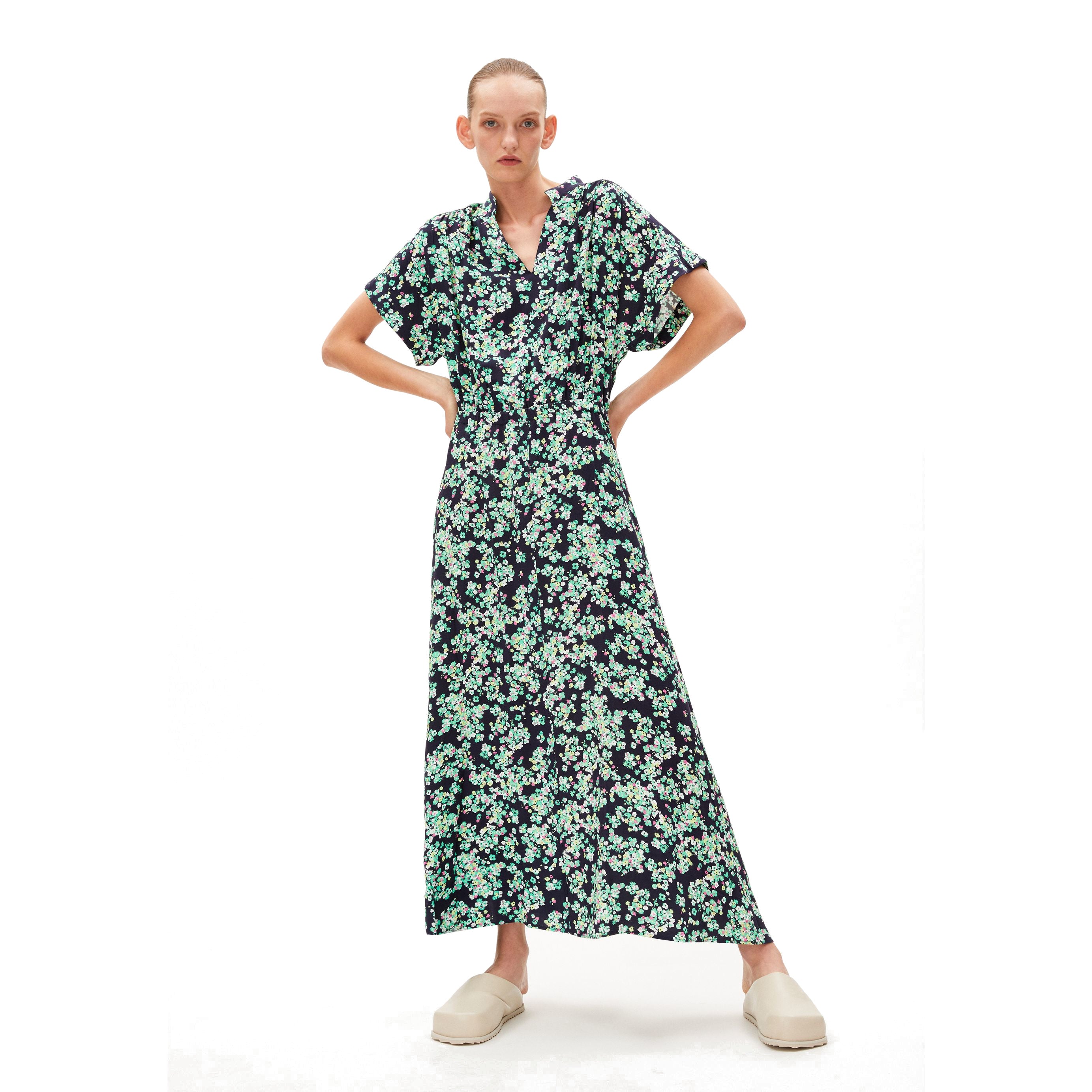 Person in a long, floral-patterned dress with short sleeves and beige shoes stands against a white background.