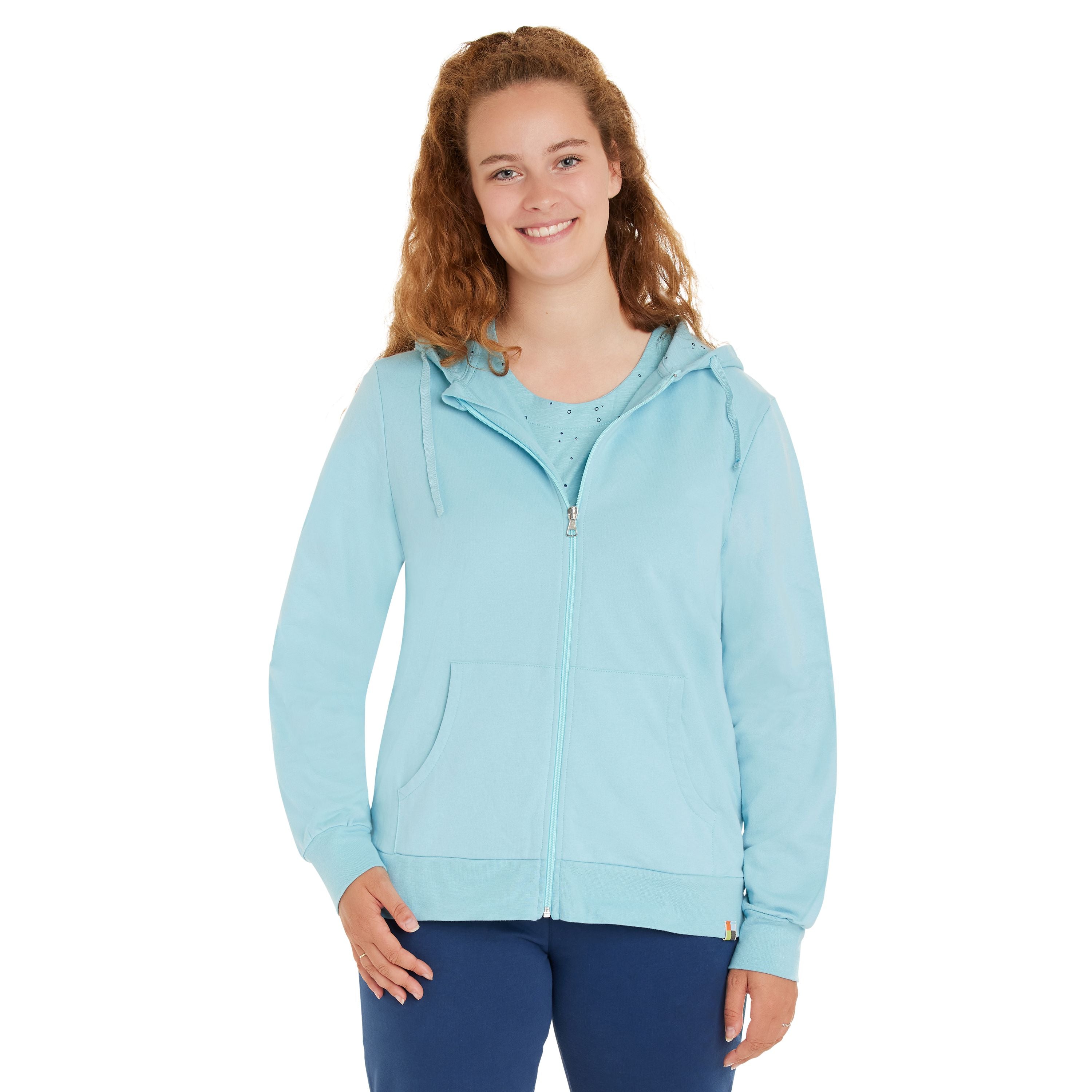 Eine junge Frau mit langem, lockigem Haar lächelt, während sie einen hellblauen Kapuzenpullover mit Reißverschluss und eine marineblaue Hose trägt und vor einem schlichten weißen Hintergrund steht.