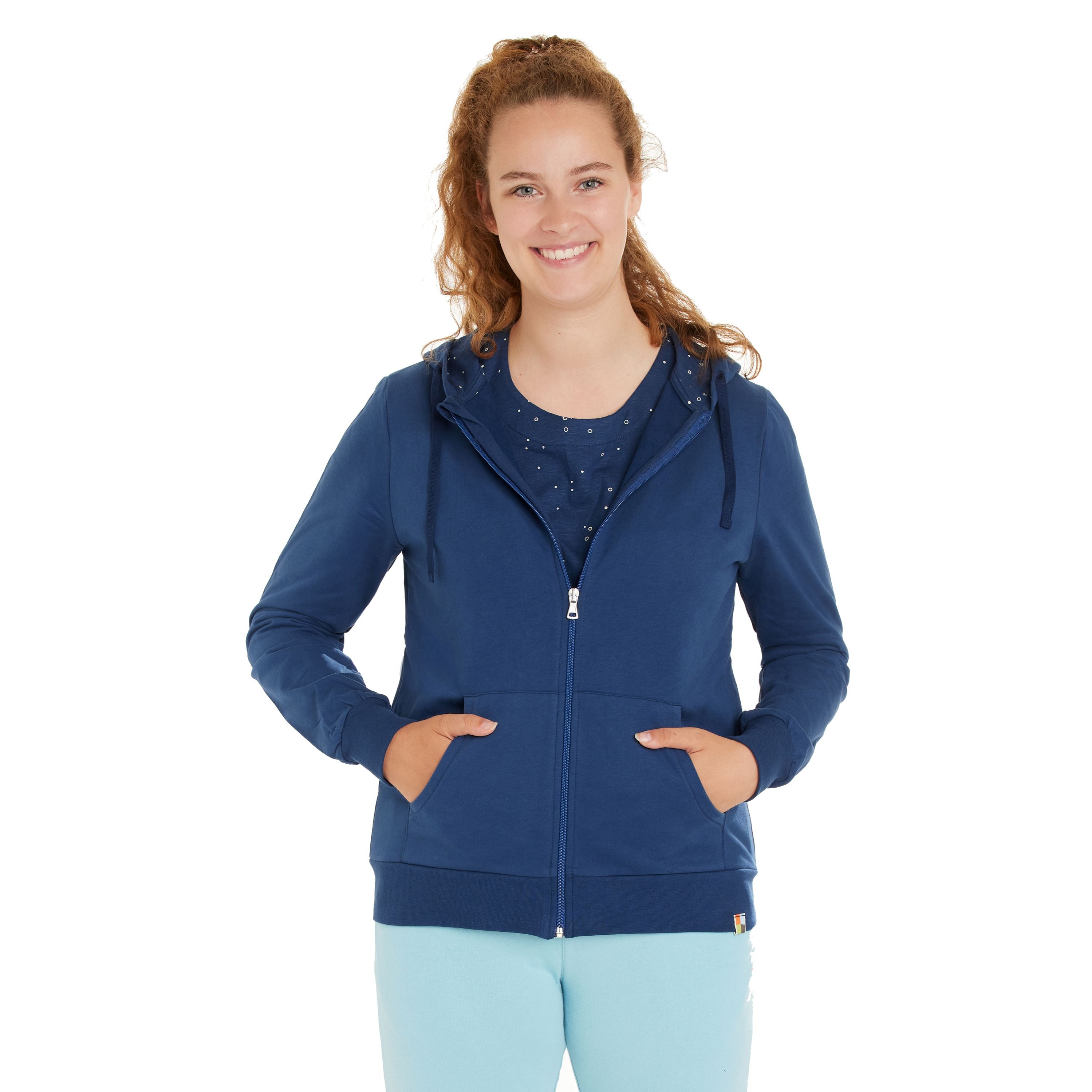 Eine junge Frau mit lockigem Haar lächelt und trägt einen blauen Kapuzenpulli mit Reißverschluss über einem passenden blauen Hemd und einer hellblauen Hose. Sie steht mit dem Gesicht nach vorne und den Händen in den Taschen des Hoodies vor einem weißen Hintergrund.