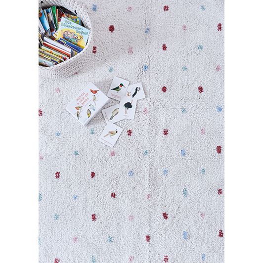 Ein strukturierter weißer Teppich mit verstreuten roten und blauen Punkten. In der oberen linken Ecke steht ein weißer Korb mit Kinderbüchern, daneben liegen ein paar illustrierte Tierkarten und ein Buch.