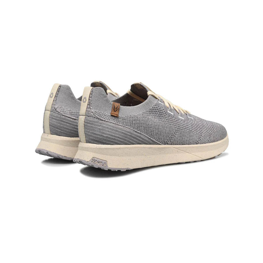 Ein Paar hellgraue, gestrickte Turnschuhe mit beigen Sohlen und Schnürsenkeln, von hinten und von der Seite gesehen, mit einem kleinen braunen Etikett in der Nähe der Ferse.