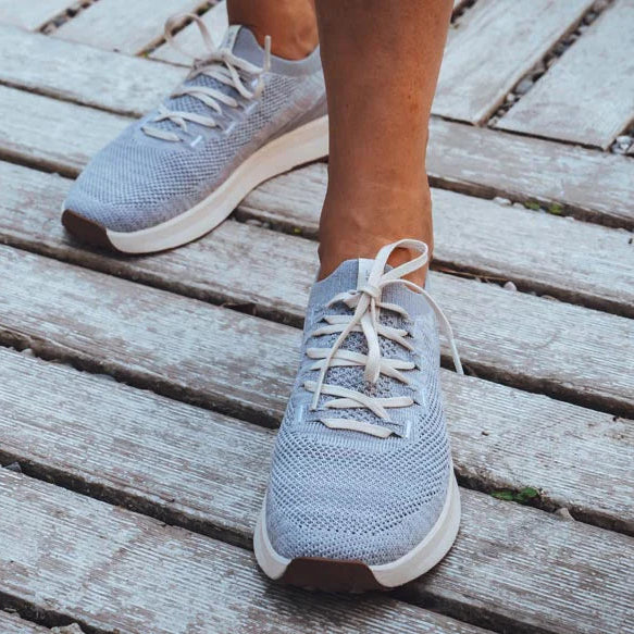 Eine Person in hellgrauen Turnschuhen mit weißen Sohlen steht auf einem verwitterten Holzdeck. Nur die Unterschenkel und Füße sind sichtbar.