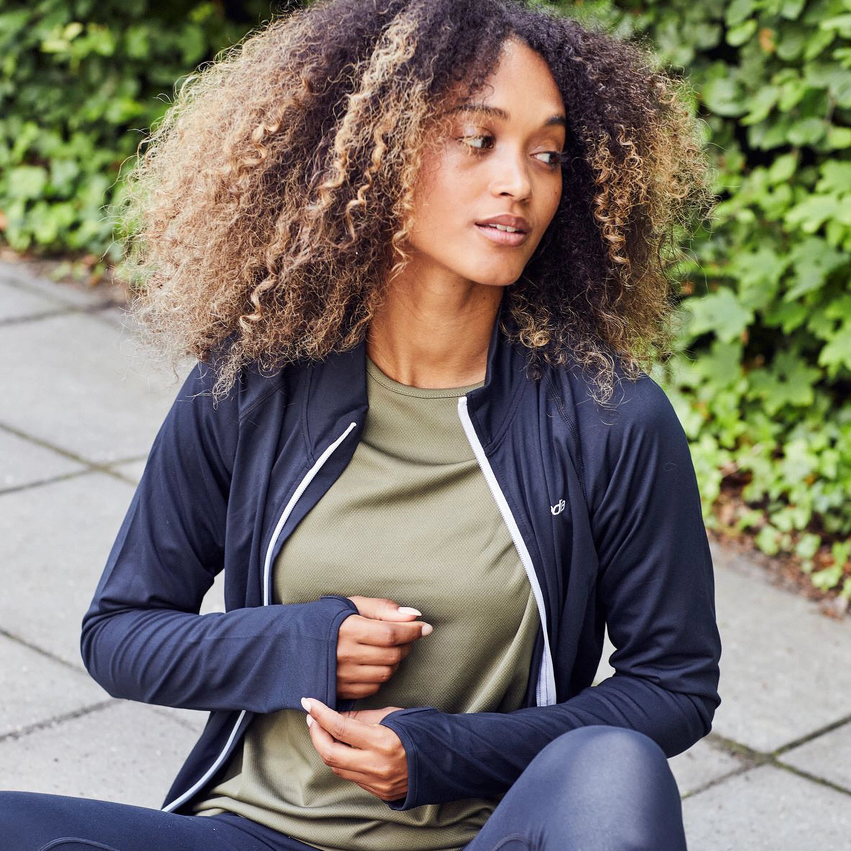 Eine Frau mit lockigem Haar sitzt auf einem Bürgersteig in Sportkleidung, darunter eine schwarze Jacke und Leggings sowie ein olivgrünes Oberteil, und blickt zur Seite, während sich im Hintergrund Grünpflanzen befinden.
