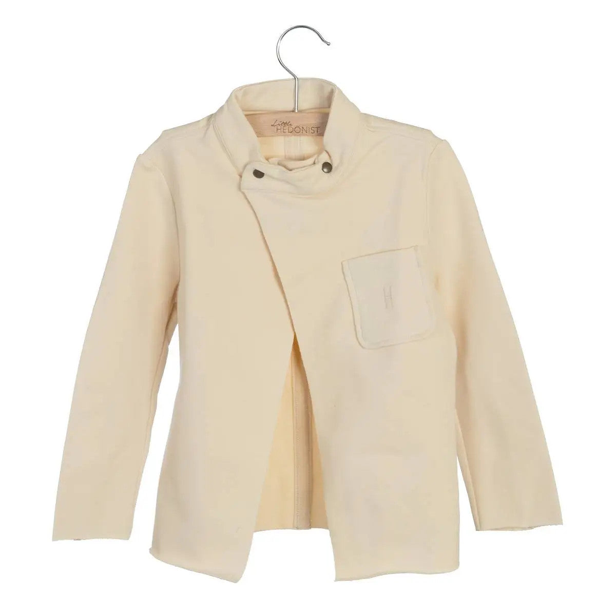 Eine hellbeige, langärmelige Jacke für Kinder mit asymmetrischer Vorderseite, Druckknöpfen am Kragen und einer einzigen Brusttasche, die auf einem Bügel vor weißem Hintergrund präsentiert wird.
