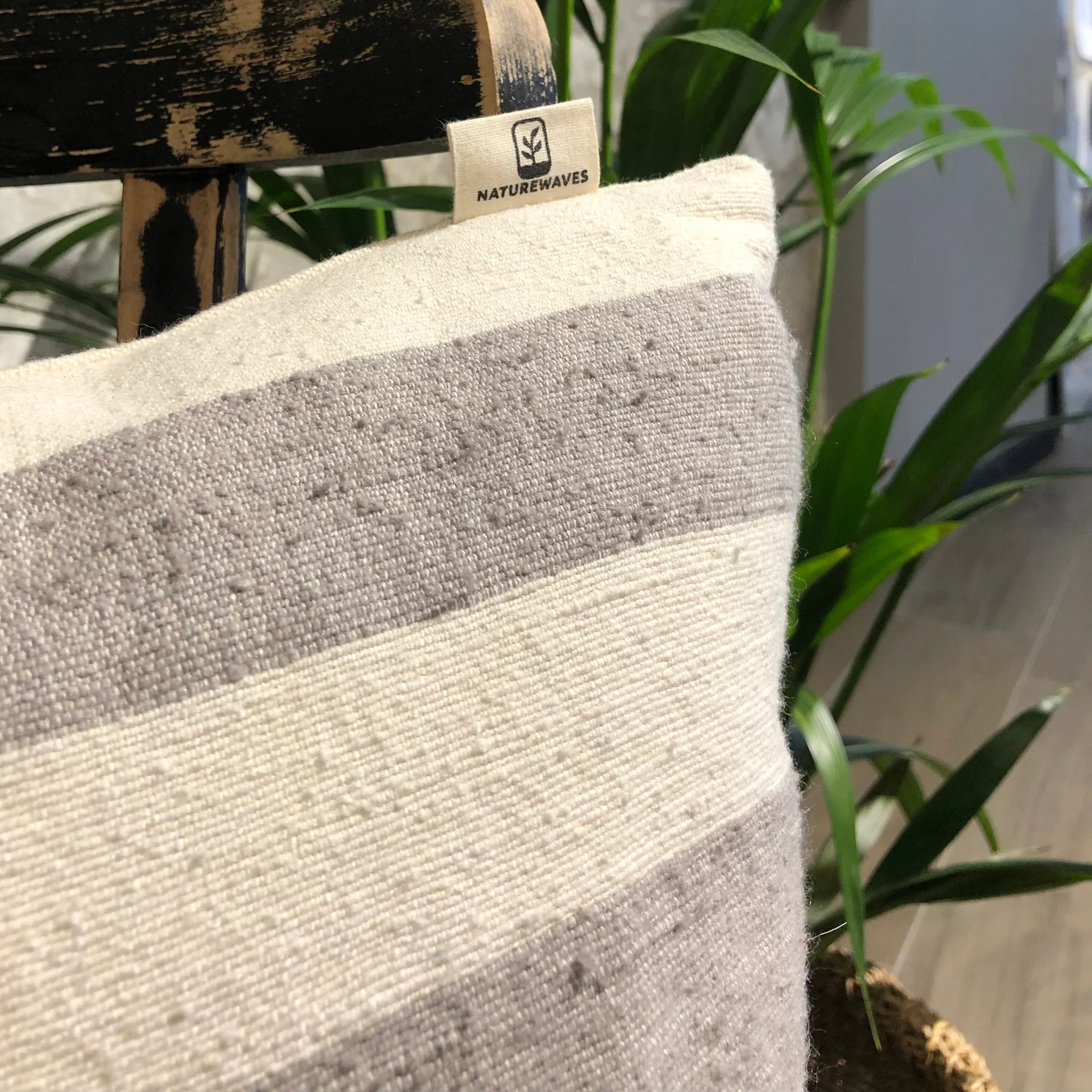 Graues und weiß gestreiftes Kissen mit "NATUREWAVES"-Etikett, auf einem Stuhl, im Hintergrund grüne Pflanzen.