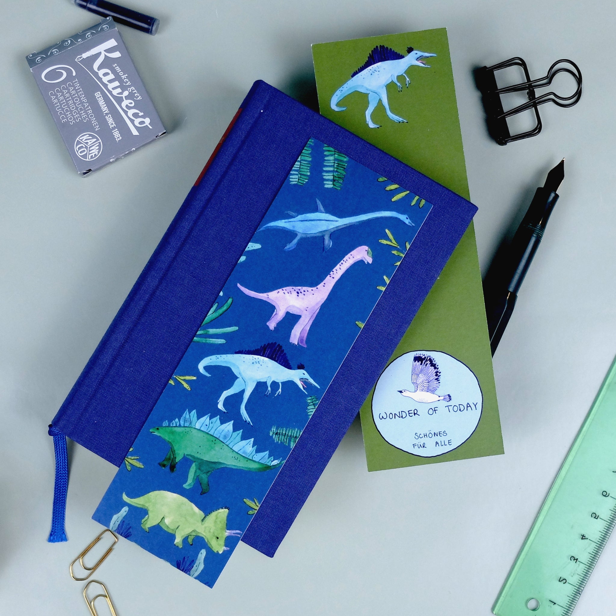 Notizbuch mit Dinosauriermotiven, daneben ein Füllfederhalter, Büroklammern, ein grünes Lineal und schwarze Tinte.