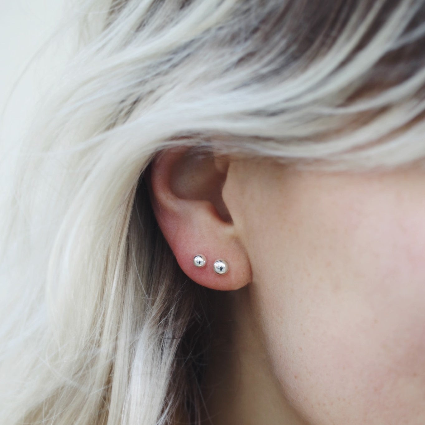Nahaufnahme eines Ohrs mit zwei kleinen, silbernen Ohrringen und blonden Haaren im Hintergrund.