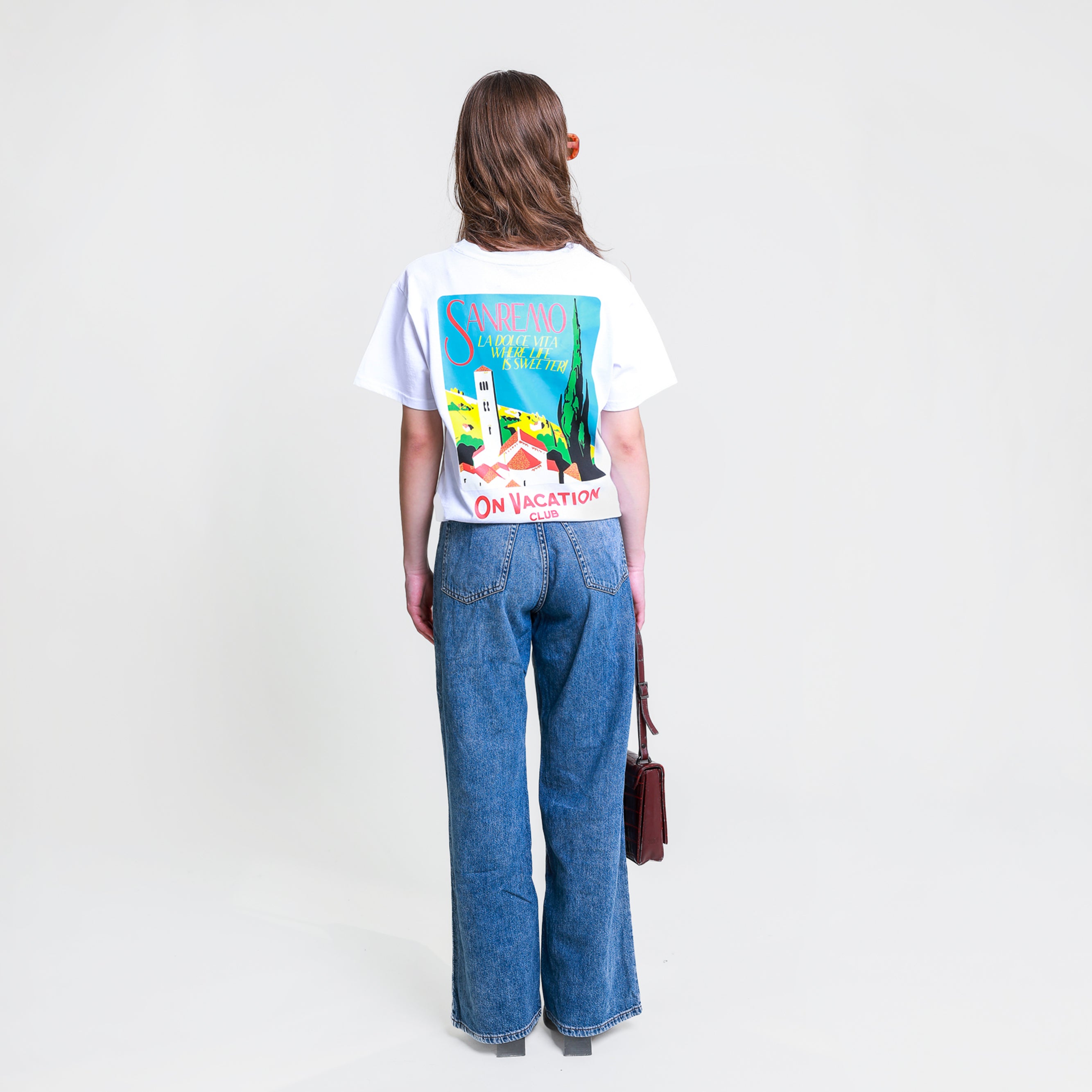 Frau mit braunem Haar trägt ein weißes T-Shirt mit Sanremo-Print und Jeans, hält eine kleine Tasche.