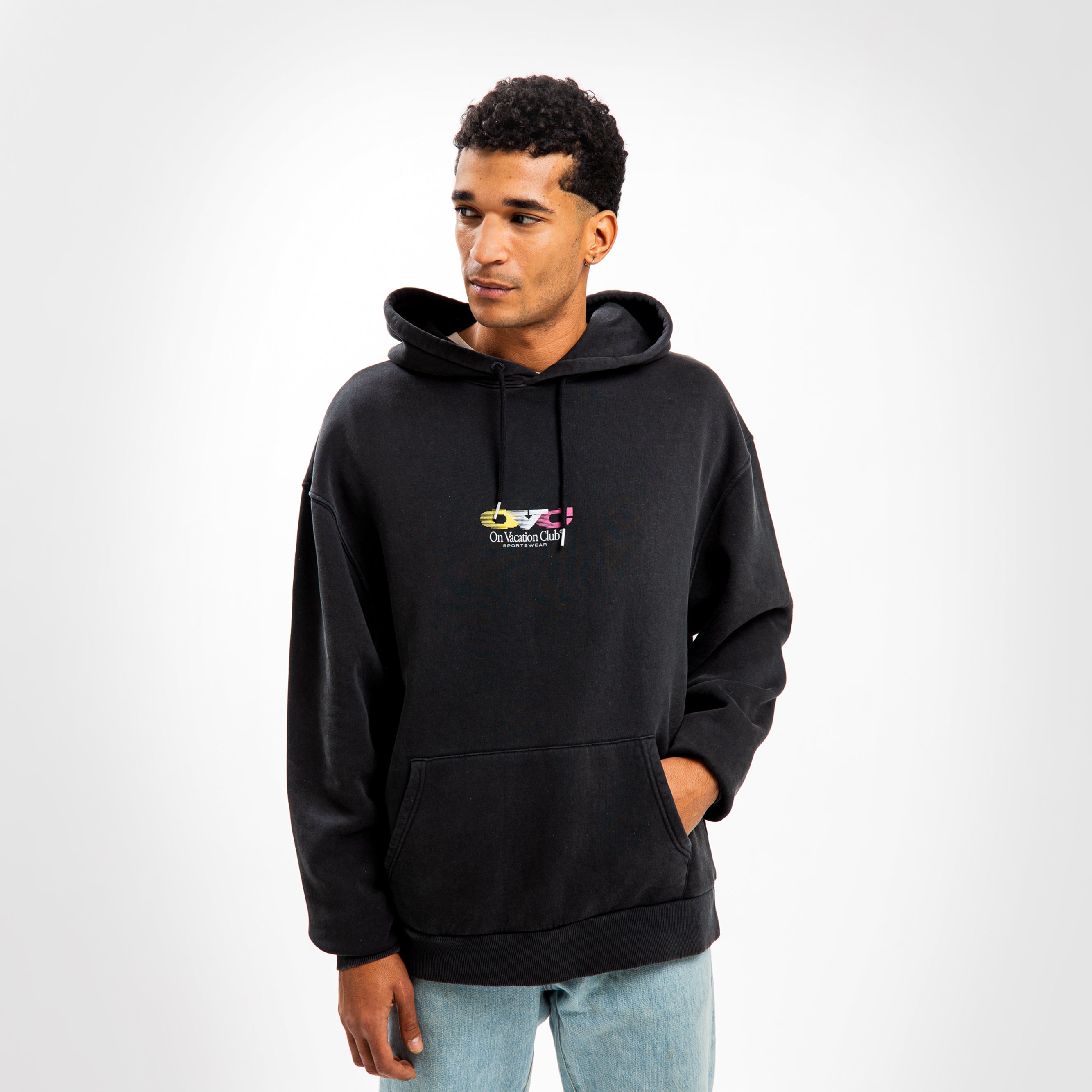Ein Mann trägt einen schwarzen Kapuzenpullover mit Aufschrift und blaue Jeans vor weißem Hintergrund.