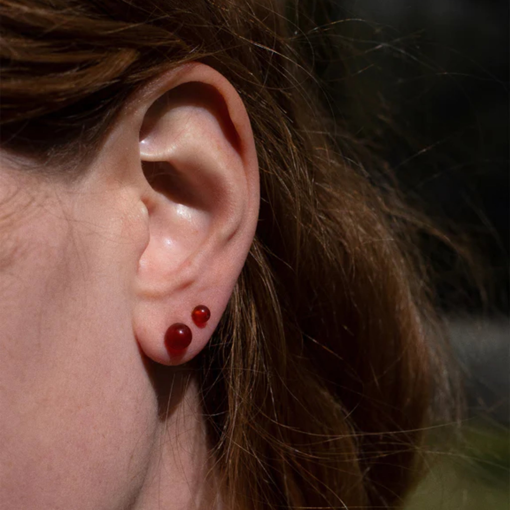 Nahaufnahme eines Ohrs mit langen braunen Haaren, das zwei rote Perlenohrringe trägt.