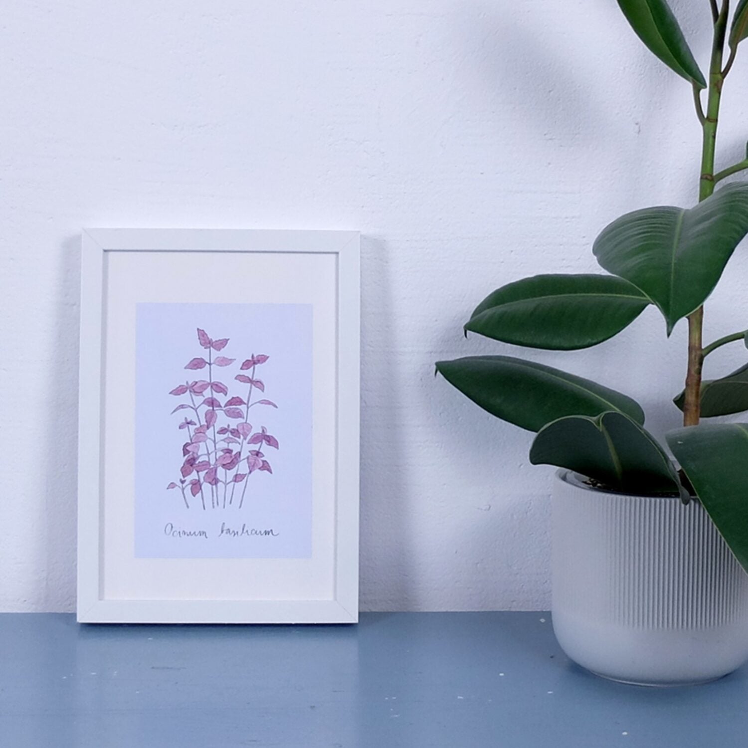 Gerahmtes Blattmuster in Rosa auf blauem Tisch neben einer Topfpflanze.