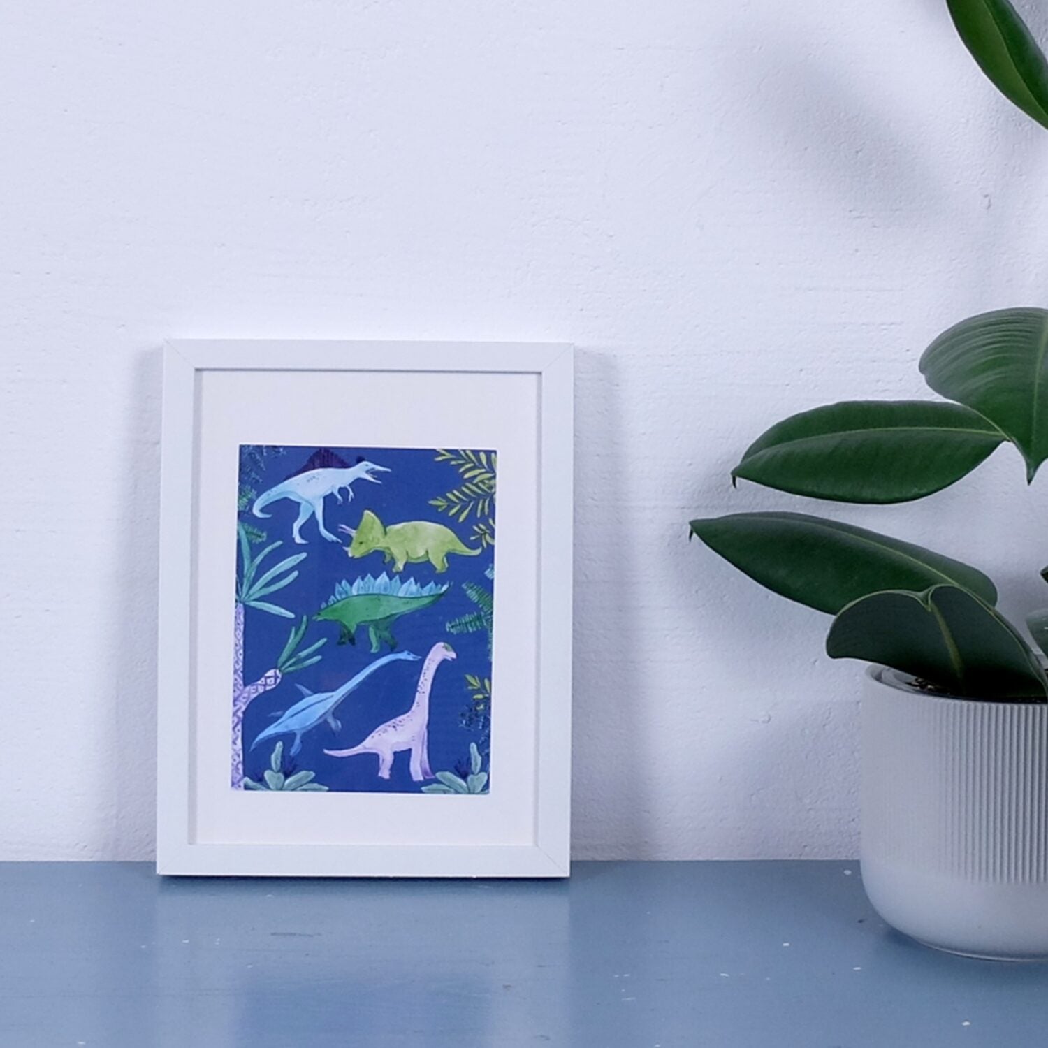 Ein gerahmtes Bild mit Dinosaurier-Illustrationen steht auf einem Tisch neben einer grünen Topfpflanze.