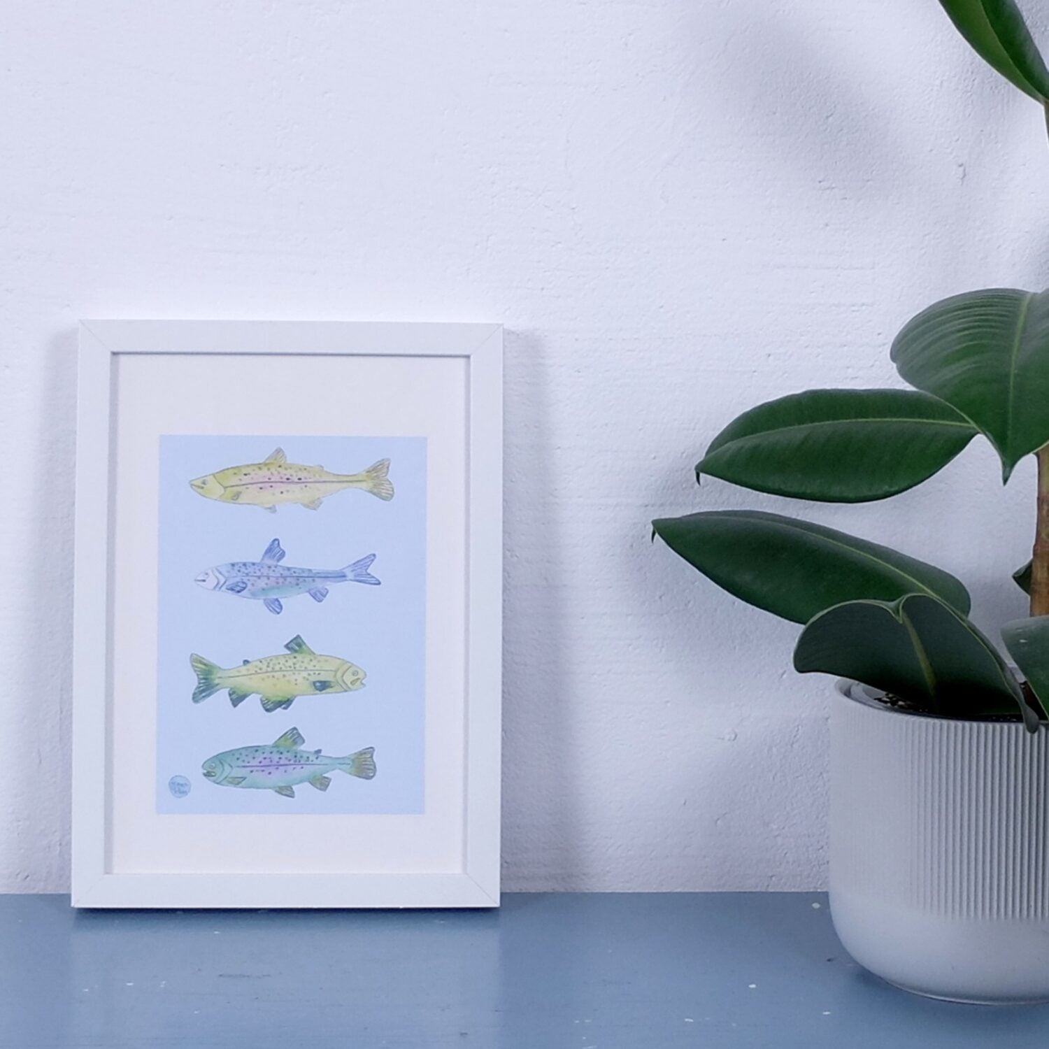 Gerahmtes Bild mit bunten Fischillustrationen neben einer Pflanze in einem weißen Topf auf einem blauen Tisch.