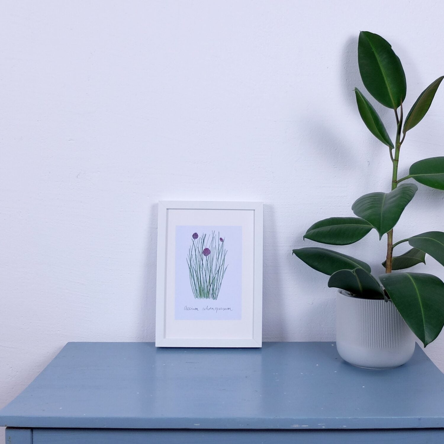 Weiße Wand mit einem gerahmten Blumenbild auf einem blauen Tisch neben einer grünen Pflanze in einem weißen Topf.