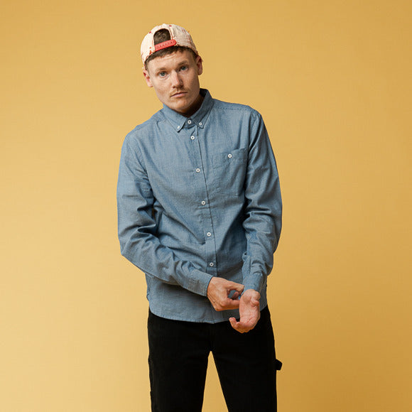 Ein Mann mit blauem Hemd und Baseballkappe steht vor einem gelben Hintergrund und richtet seinen Ärmel.