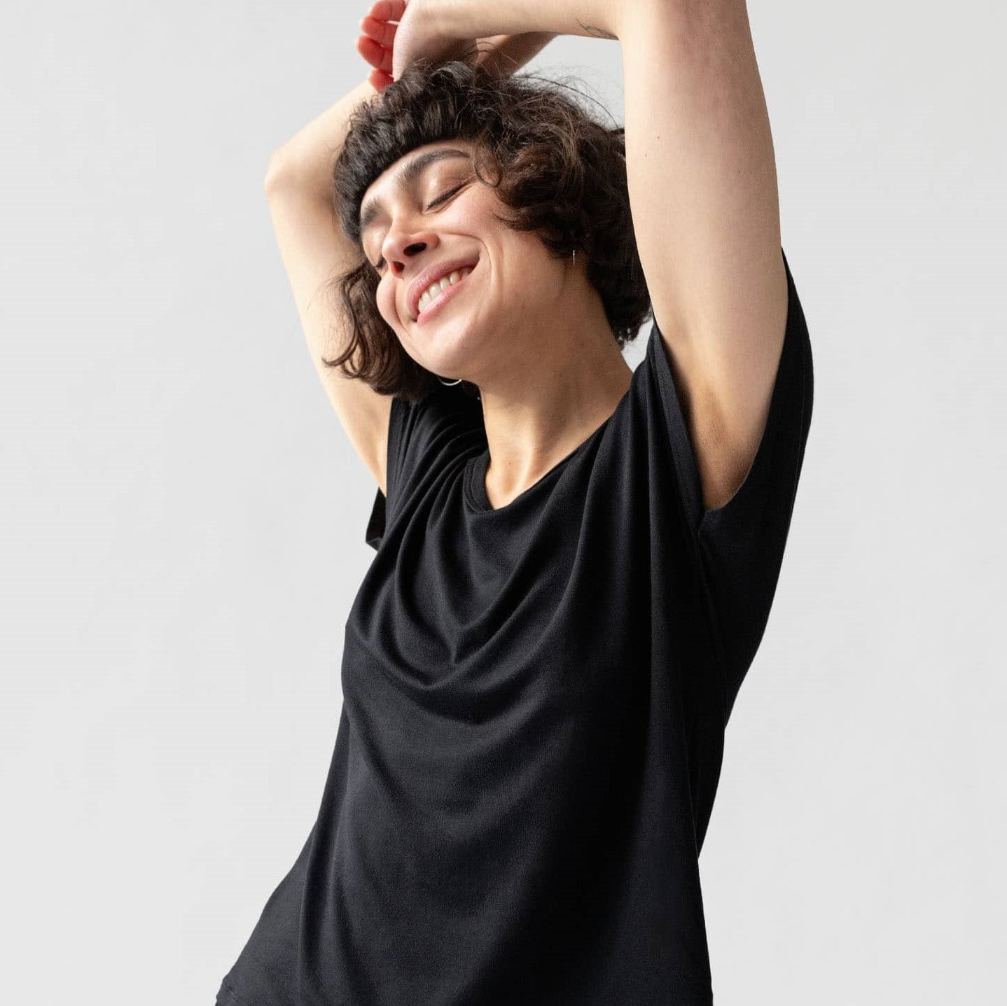 Person mit lockigem Haar in schwarzem T-Shirt lächelt mit geschlossenen Augen und erhobenen Armen.