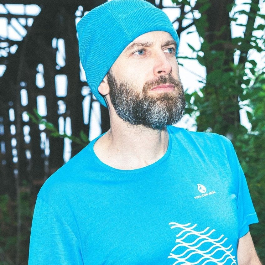Ein Mann mit Bart trägt ein blaues T-Shirt und eine blaue Mütze, umgeben von Bäumen.