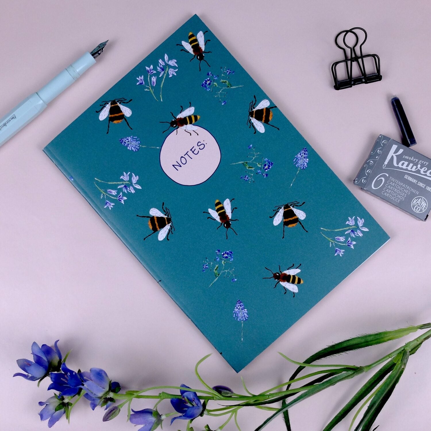 Notizbuch mit Bienen- und Blumenmotiven, umgeben von einem Stift, Klammer und lila Blumen auf rosa Hintergrund.